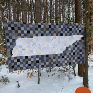 Könnte beinhalten: Ein rechteckiger Quilt mit einer verpixelten Karte von Tennessee in hellen Creme- und Beigetönen, umgeben von einem dunkelgrauen und schwarzen Karomuster. Der Text lautet: TENNESSEE: A Pixelated State Quilt. Fertige Größe: 196,85 cm x 107,95 cm.
