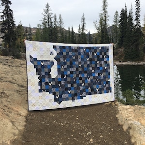 Puede incluir: Una colcha azul y blanca con la forma del estado de Washington, con un estampado de ciervo. La colcha se exhibe al aire libre con un fondo de árboles y un lago.