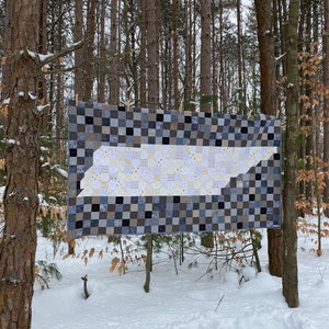 May include: A handmade quilt with a Tennessee state outline design. The quilt features a patchwork pattern in shades of gray, black, and white. It is displayed outdoors, hanging between trees in a snowy environment.