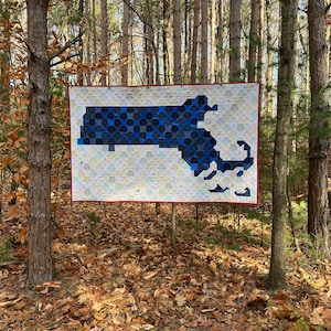 May include: A blue and white quilt depicting the state of Massachusetts hanging in a woodland setting. The quilt is made of patchwork squares and is surrounded by trees.