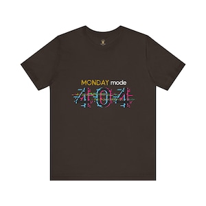 Może przedstawiać: Ciemnobrązowy t-shirt z żółtym napisem "MONDAY mode" i kolorowym wzorem glitch z napisem "404" w kolorze niebieskim, różowym i zielonym. Koszulka ma okrągły dekolt i krótkie rękawy.