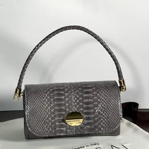 Puede incluir: Bolso de mano gris de piel de serpiente con asa curva y herrajes dorados. El bolso rectangular presenta un cierre de solapa con un acento dorado circular. El bolso está sobre una tela blanca con el texto "UNICA BAG LAB" y "Handmade in Italy".
