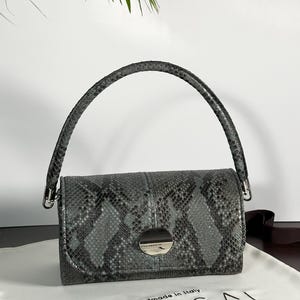Puede incluir: Bolso rectangular gris con estampado de piel de serpiente, asa curva y cierre plateado. El bolso lleva la etiqueta "UNICA BAG LAB" y "Handmade in Italy" sobre una tela blanca.