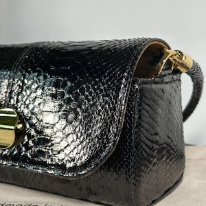 Pode incluir: Bolsa retangular preta com um padrão texturizado semelhante ao de um réptil. A bolsa apresenta um fecho e uma fixação de alça dourados. As palavras "handmade in Italy" são visíveis na superfície inferior.