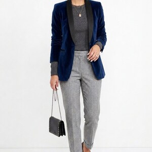 May include: A navy blue velvet blazer with a black lapel is worn over a gray sweater. The outfit includes gray pinstripe pants and a black purse with a chain strap. The ensemble is a stylish, professional look.
