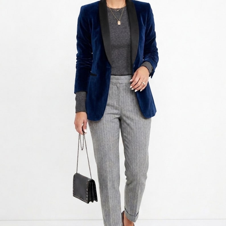May include: A navy blue velvet blazer with a black lapel is worn over a gray sweater. The outfit includes gray pinstripe pants and a black purse with a chain strap. The ensemble is a stylish, professional look.