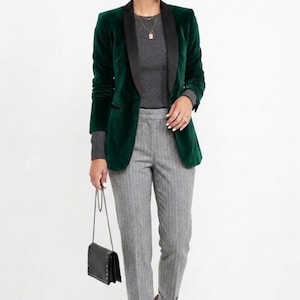 May include: A dark green velvet blazer with a black lapel, paired with a gray long-sleeve top and gray pinstripe pants. A small black purse with a chain strap is held in the hand.