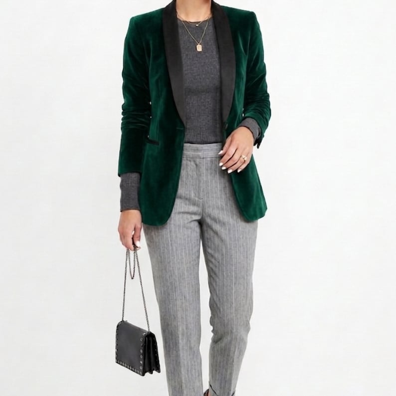 May include: A dark green velvet blazer with a black lapel, paired with a gray long-sleeve top and gray pinstripe pants. A small black purse with a chain strap is held in the hand.