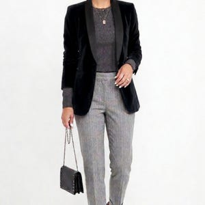 May include: A black velvet blazer with a satin lapel is paired with a gray ribbed sweater and gray pinstripe trousers. A small black purse with a chain strap completes the outfit. The ensemble is suitable for business or formal occasions.