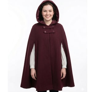 May include: A burgundy hooded cape with two button closures. The cape has a satin lining in the hood and is worn over a white long-sleeved shirt. The cape is a stylish outerwear garment.