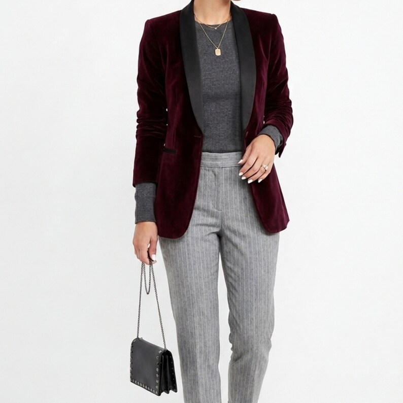 May include: A burgundy velvet blazer with a black satin lapel is worn over a gray ribbed long-sleeve shirt. The outfit includes gray pinstripe trousers and a black studded purse with a chain strap. The model is wearing gold necklaces.