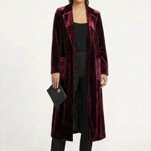 Peut inclure: Un long manteau en velours bordeaux avec un col à revers classique et deux poches avant. Le manteau est porté sur un haut noir et un pantalon noir. Une pochette noire est tenue à la main.