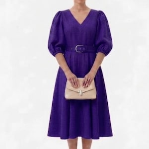 以下が含まれることがあります： Vネックと短いパフスリーブの深い紫色のドレス。 ドレスは、ウエストを締めるお揃いのベルトとベージュのクラッチが特徴です。 ドレスはフレアスカートで、テクスチャード加工された生地で作られています。