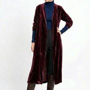 May include: A long-sleeved, open-front duster cardigan made of deep burgundy velvet fabric. The garment features a straight, floor-length silhouette with long sleeves and no visible buttons or closures, layered over a dark blue turtleneck top.