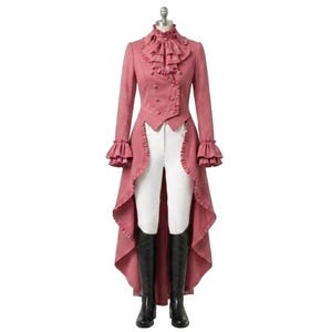 Puede incluir: Un frac rosa con cuello alto con volantes, doble botonadura y mangas largas con puños con volantes. El abrigo tiene una espalda larga y fluida y se combina con pantalones blancos y botas de montar negras. El atuendo se exhibe en un maniquí.