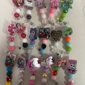 May include: A collection of beaded badge reels with various decorative toppers. Designs include a Marilyn Monroe heart, a coffee cup, Hello Kitty, bows, a cross, Betty Boop, a ghost, the word "MOM", a heart with "Jesus loves you", a van, a video game character, a fairy, a unicorn, and a Jack Skellington design.
