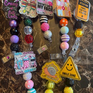Puede incluir: Una colección de carretes de insignia con cuentas con varios diseños y texto. Los carretes presentan frases como "Crazy Cat Lady", "If my face doesn't say it" y "I just work here". Las cuentas son de varios colores, incluyendo negro, morado, rosa y amarillo.