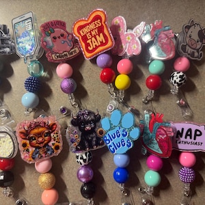 May include: A collection of badge reels with various designs. The designs include a football helmet, an IV bag, a cute cartoon character, a slice of toast, a butterfly, a heart, a cow, a pill, a cow with sunflowers, a highland cow, a paw print, and a pillow with the text "NAP ENTHUSIAST".