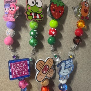 Puede incluir: Una colección de carretes retráctiles para insignias con varios diseños. Los carretes presentan personajes de dibujos animados, una fresa y los textos "Back & Body Hurts" y "Silly Goose". Cada carrete tiene un acento de cuentas en varios colores.