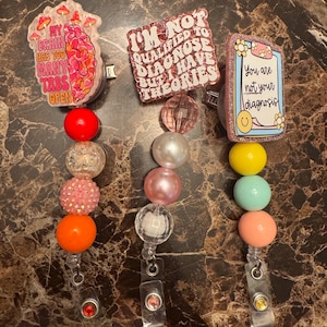 May include: Three retractable badge holders with colorful beaded accents. One has a brain graphic with text "MY BRAIN HAS TOO MANY TABS OPEN". Another says "I'M NOT QUALIFIED TO DIAGNOSE BUT HAVE THEORIES". The third says "You are not your diagnosis".