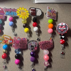 Puede incluir: Una colección de carretes de insignia con cuentas con varios diseños y texto. Los carretes presentan frases como "Medical Assistant", "Soon to be Nurse" y "Future LPN". Otros diseños incluyen un girasol, una pastilla, un oso de peluche y un corazón de enfermera.