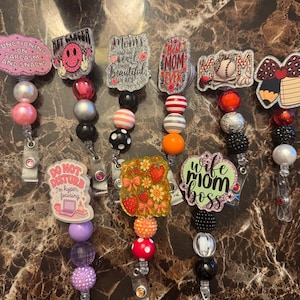 Puede incluir: Una colección de carretes de insignias con cuentas con varios diseños y texto. Los carretes presentan frases como "Functioning on Sarcasm and Snacks", "Hey Gangster" y "Wife Mom Boss". Cada carrete tiene un diseño único, incluyendo fresas, caras sonrientes y béisbol.