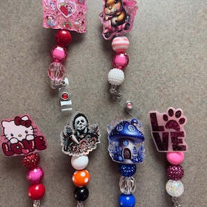 Puede incluir: Una colección de carretes de insignia decorativos con varios diseños. Los carretes presentan imágenes de una fresa, una ardilla en un scooter, Hello Kitty con la palabra "LOVE", un personaje de película de terror, una casa de hongos y una huella de pata con la palabra "LOVE". Cada carrete tiene una cadena de cuentas.