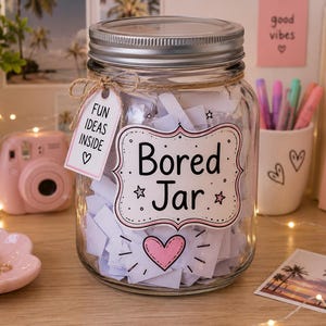 K&ouml;nnte beinhalten: Ein klares Glas mit silbernem Deckel, gef&uuml;llt mit wei&szlig;en Papierstreifen. Das Glas hat ein Etikett mit der Aufschrift "Bored Jar" mit einem rosa Herz. Ein Anh&auml;nger lautet "Fun Ideas Inside". Eine rosa Kamera und eine rosa Schale mit goldenen Ringen sind in der N&auml;he.
