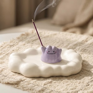 Puede incluir: Un porta incienso de cerámica con un personaje morado sonriente, colocado sobre un plato blanco con forma de nube. Un palito de incienso encendido emite humo. El soporte está colocado sobre una superficie beige texturizada.