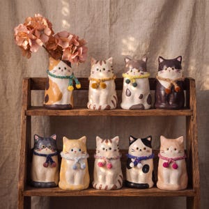 Puede incluir: Una colección de nueve jarrones de cerámica con forma de gato dispuestos en un estante de madera. Cada figura de gato presenta un patrón de pelaje único, detalles faciales pintados y una pequeña bufanda tejida a mano con pompones alrededor del cuello. Un jarrón contiene flores rosas.