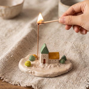 Puede incluir: Un porta-incienso de cerámica con forma de pequeña casa con techo verde y lateral amarillo, sobre una base beige texturizada con pequeños árboles verdes y amarillos. Una varilla de incienso fina y encendida se alza junto a la casa.