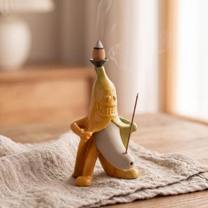 May include: A ceramic incense burner shaped like a yellow anthropomorphic banana with a grinning face. The figure holds a thin incense stick in one hand and features a small platform on its head to support a burning cone, emitting wisps of smoke.