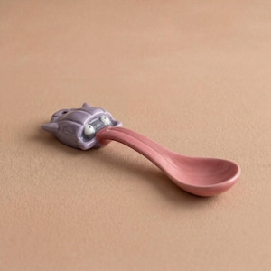 May include: A novelty ceramic spoon featuring a purple monster head with a pink tongue forming the spoon's bowl. The monster has white eyes and an open mouth. The spoon is set against a neutral background.