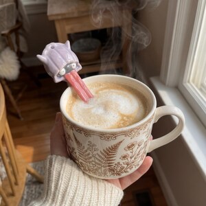 May include: A ceramic mug filled with coffee, with a novelty spoon shaped like a lavender monster with a pink tongue. The mug has a brown and white floral design. Steam rises from the coffee.
