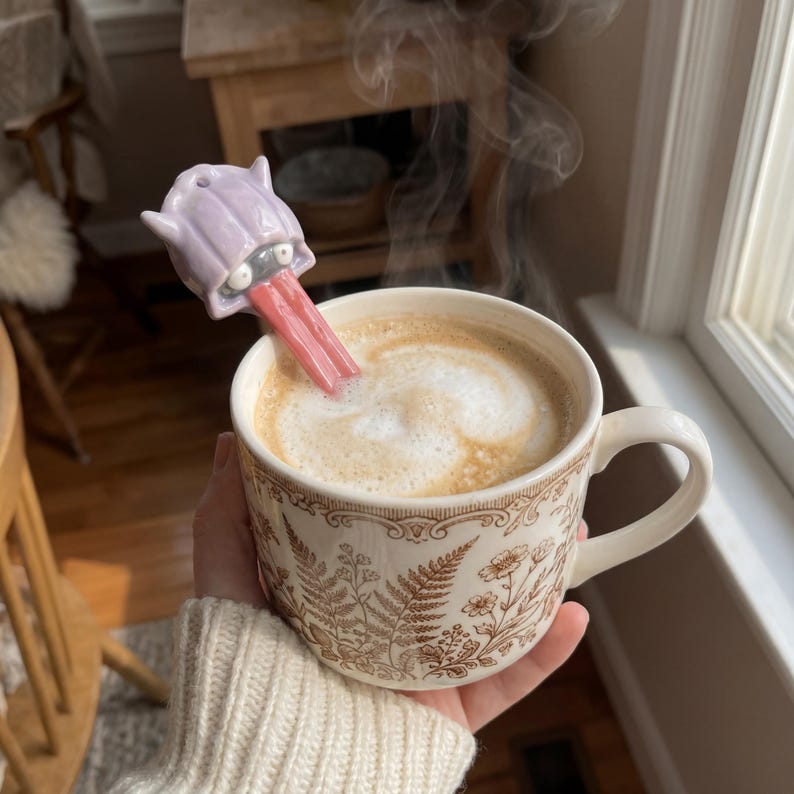 May include: A ceramic mug filled with coffee, with a novelty spoon shaped like a lavender monster with a pink tongue. The mug has a brown and white floral design. Steam rises from the coffee.