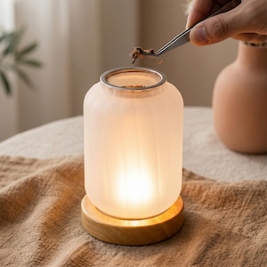 May include: A cylindrical frosted glass wax warmer with vertical ribbing, illuminated from within, resting on a circular light-colored wooden base. The top features a silver-toned metal mesh insert for holding wax melts or aromatic botanical materials.