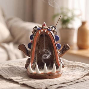 May include: A brown ceramic incense burner shaped like a creature with a wide-open mouth, sharp white teeth, and blue spiral accents. A lit incense cone sits on top, releasing a thin trail of smoke that flows downward into the creature's mouth.
