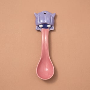 May include: A whimsical ceramic spoon with a cartoonish face design. The spoon features a lavender head with blue eyes and a pink tongue extending down to the pink spoon bowl. A hole is at the top for hanging.