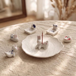 Peut inclure: Un porte-encens en céramique blanche en forme de lapin avec des motifs floraux roses, posé sur une assiette blanche texturée. Autour de l'assiette se trouvent cinq petites figurines d'animaux en céramique, dont un chat et un chien.