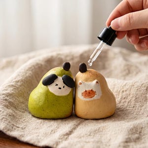 Peut inclure: Deux petites figurines en céramique en forme de poire avec des visages d'animaux peints. L'une est verte avec un visage de chien aux oreilles noires, l'autre est beige avec un visage de chat blanc. Un compte-gouttes en verre est au-dessus.