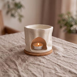 Puede incluir: Un calentador de cera de cerámica color crema con acabado moteado y textura irregular arrugada. El recipiente presenta una amplia abertura arqueada en la base que contiene una pequeña vela de té encendida, sobre un posavasos circular de corcho.