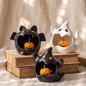 May include: Three ceramic Halloween figurines featuring a black bat, a white ghost, and a black witch hat, each with a wide open mouth containing a small orange jack-o'-lantern. The figures have yellow or black painted eyes and smooth surfaces.