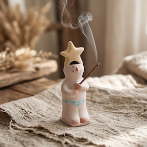 Könnte beinhalten: Ein Räucherstäbchenhalter aus Keramik in Form einer Figur mit einem gelben sternförmigen Hut und einer hellblauen Unterhose mit Punkten. Die Figur hat eine kleine rote Nase und schwarze Augen und hält ein braunes Räucherstäbchen mit weißem Rauch.