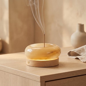May include: An amber-toned incense holder with a wooden base. A lit incense stick emits smoke. The holder has a rounded shape and sits on a light-coloured wooden surface. A neutral-toned vase is in the background.