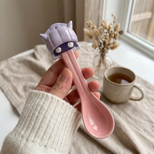 May include: A pink ceramic spoon with a cartoon character head on the handle. The character has a purple head, a dark purple mask, and white eyes. The spoon is held in a hand.