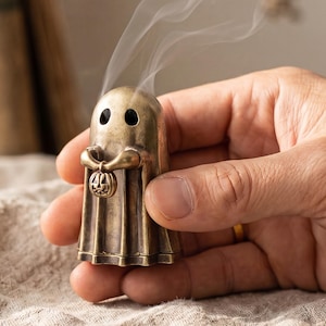 May include: A small, antique-style brass ghost figurine incense burner with a weathered finish. The ghost has two dark eye cutouts and holds a miniature carved pumpkin in its arms. Wisps of white smoke rise from the top of the ghost's head.