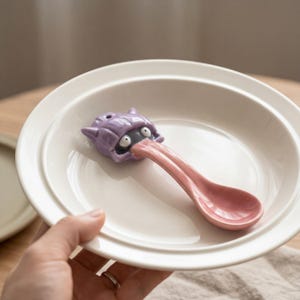 May include: A ceramic spoon featuring a purple monster head. The monster's pink tongue extends to form the spoon's handle and bowl. The spoon is placed on a white plate.