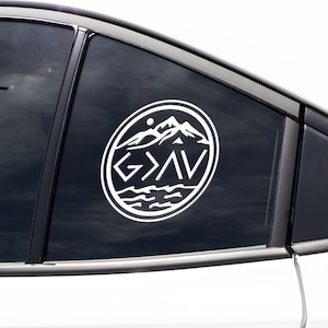 May include: White vinyl car sticker with a circular design. The design features a mountain range, sun, and water, with the text "G > AV". The sticker is applied to a car window.
