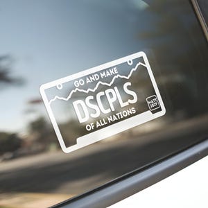 May include: White rectangular sticker with black text on a car window. The sticker reads "GO AND MAKE DSCPLS OF ALL NATIONS" with a mountain range graphic. A smaller box in the corner has the text "MATT 28:19".
