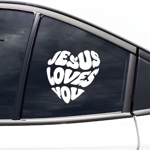 Può includere: Adesivo per auto in vinile bianco a forma di cuore con la scritta "JESUS LOVES YOU" in un carattere grassetto e arrotondato. L'adesivo è applicato sul finestrino di un'auto.
